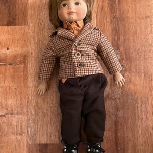 Könnte beinhalten: Eine Porzellanpuppe mit braunen Haaren, die eine braune und weiße karierte Jacke, ein braunes Hemd, eine braune Hose und schwarze Schuhe trägt.