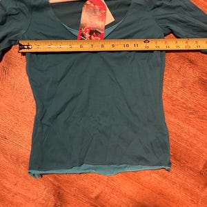 May include: Teal long-sleeve shirt with a scoop neck and a layered design. The shirt is on a hanger and has a tag that says "energe."