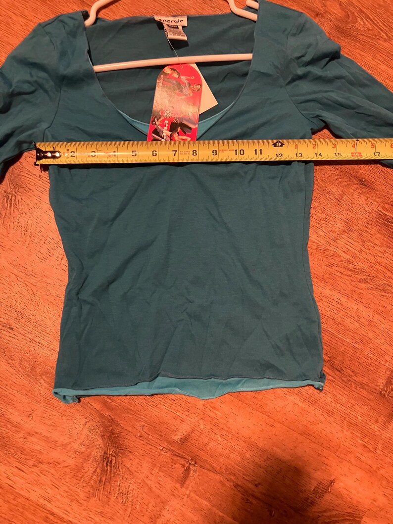 May include: Teal long-sleeve shirt with a scoop neck and a layered design. The shirt is on a hanger and has a tag that says "energe."