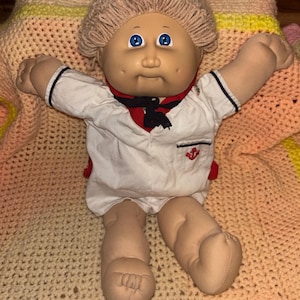 May include: A vintage Cabbage Patch Kid doll, featuring a soft, fabric body and a vinyl head with blue eyes and a neutral expression. The doll is dressed in a white sailor outfit with red accents and a black necktie. The doll is posed with arms outstretched.