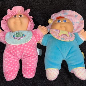 May include: Two vintage Cabbage Patch Kids dolls. One doll is wearing a pink polka-dot outfit and matching bonnet. The other doll is wearing a blue outfit and a pink bonnet. Both dolls have bibs that say "Babyland".