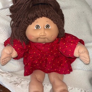 Peut inclure: Une poupée au corps en tissu souple, au teint clair et aux cheveux en laine brune attachés par un élastique noir. La poupée a des yeux marron, des joues avec des fossettes et porte une robe rouge à manches longues à motifs floraux.