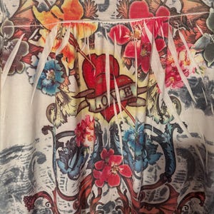 Puede incluir: Top color crema con un diseño floral vibrante. El diseño incluye un corazón rojo con la palabra "LOVE" en una pancarta, rodeado de flores coloridas y adornos ornamentados. El top tiene un escote fruncido.