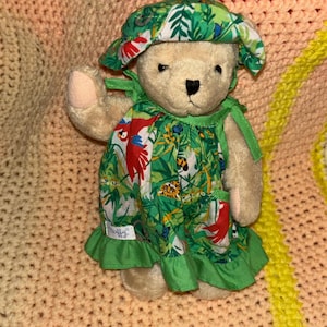 Può includere: Un orsacchiotto beige con un vestito verde con stampa tropicale e un cappello abbinato. Il vestito presenta un vivace motivo di pappagalli, foglie e altri elementi della giungla. Il cappello ha un bordo bianco. L'orso alza una zampa.