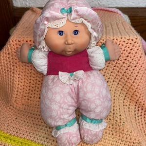 May include: A vintage baby doll with blue eyes, wearing a pink floral print outfit with a matching bonnet. The doll's outfit includes a pink ribbed top, and the ensemble is accented with teal bows and white lace trim. The doll is resting on a peach-colored blanket.