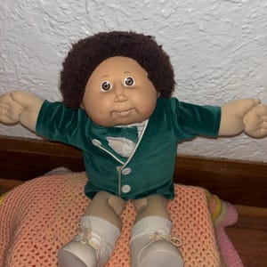 May include: A vintage Cabbage Patch Kid doll. The doll has a tan complexion, brown curly hair, and large, expressive eyes. It wears a green velvet jacket, white shirt, and white shoes. The doll is seated on a peach-colored crocheted blanket.