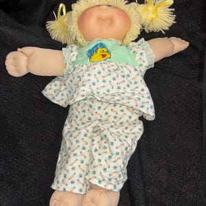K&ouml;nnte beinhalten: Eine Vintage-Puppe mit blonden Wollhaaren, die zu Z&ouml;pfen gestylt sind. Die Puppe tr&auml;gt ein hellgr&uuml;nes Oberteil mit einem gelben Fischmuster und ein Outfit mit floralem Muster. Die Arme der Puppe sind ausgestreckt.