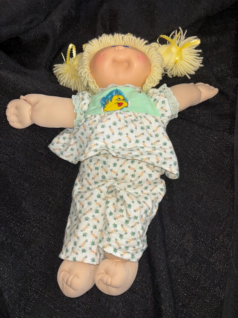 K&ouml;nnte beinhalten: Eine Vintage-Puppe mit blonden Wollhaaren, die zu Z&ouml;pfen gestylt sind. Die Puppe tr&auml;gt ein hellgr&uuml;nes Oberteil mit einem gelben Fischmuster und ein Outfit mit floralem Muster. Die Arme der Puppe sind ausgestreckt.