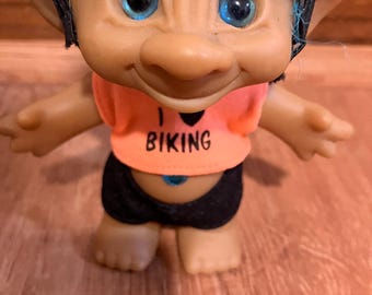 May include: A troll doll wearing a yellow and black baseball cap, an orange shirt with the text "I ♥ BIKING", and black shorts. The troll has blue eyes and brown hair.
