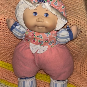 Peut inclure: Une poupée vintage au corps doux et en peluche. La poupée a un visage rond avec des yeux bleus et une expression douce. Elle porte un bonnet fleuri, une robe rose et fleurie et un pantalon rose. Les bras et les jambes de la poupée sont doux et rembourrés, avec des accents rayés bleus et blancs.