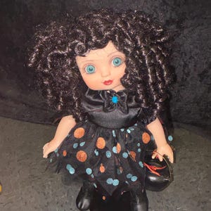 May include: A doll with curly black hair, blue eyes, and red lips. The doll wears a black dress with orange and blue polka dots, a black bow, and holds a small black cauldron. The doll is a Halloween-themed decorative item.
