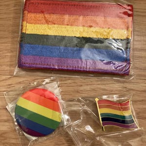 Peut inclure: Un portefeuille à rayures arc-en-ciel, un bouton rond et une épingle drapeau, tous dans des emballages individuels. Le portefeuille présente des rayures horizontales rouges, oranges, jaunes, vertes, bleues et violettes.