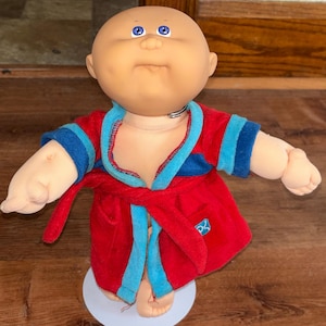 Peut inclure: Poupée Cabbage Patch Kid vintage vêtue d'un peignoir rouge avec des bordures bleues. La poupée a une tête chauve, des yeux bleus et une expression neutre. La robe est nouée à la taille et porte un petit emblème.