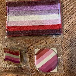 Peut inclure: Trois articles représentant le drapeau de la fierté lesbienne : un patch en tissu, une épingle et un bouton. Le patch est un morceau de tissu rectangulaire avec les couleurs du drapeau de la fierté lesbienne en rayures. L’épingle est une épingle dorée avec les couleurs du drapeau de la fierté lesbienne en rayures. Le bouton est un bouton rond avec les couleurs du drapeau de la fierté lesbienne en rayures.