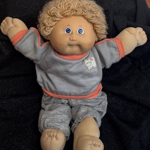 Peut inclure: Poupée vintage avec un corps en tissu doux et un visage en vinyle. La poupée a des cheveux blonds bouclés, des yeux bleus et porte un haut gris avec une bordure orange et un pantalon assorti. Un petit chat est sur le haut.