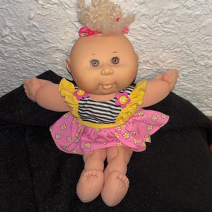 Peut inclure: Une poupée vintage avec une tête chauve, une peau claire et des yeux bruns. La poupée porte un haut rayé noir et blanc avec des manches à volants jaunes et une jupe à fleurs roses. Ses cheveux blonds sont coiffés d'un nœud rose.