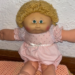 Könnte beinhalten: Eine Vintage-Puppe mit einem Vinylgesicht, grünen Augen und einer blonden Garnfrisur. Die Puppe trägt ein rosa Kleid mit Blumenmuster, Spitzensaum und passendem Strampler. Sie sitzt auf einer rosa gehäkelten Decke.
