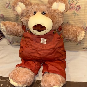 Può includere: Un orsacchiotto di peluche con pelliccia marrone chiaro, orecchie interne color crema e un muso liscio di colore chiaro con un naso marrone scuro. L'orso indossa una salopette in tessuto color ruggine con una piccola etichetta rettangolare.