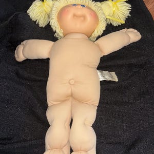 K&ouml;nnte beinhalten: Eine Vintage Cabbage Patch Kid Puppe mit einem weichen Stoffk&ouml;rper und einem Vinylkopf. Die Puppe hat blonde Zopf-Z&ouml;pfe, die mit gelben B&auml;ndern gebunden sind, blaue Augen und rosige Wangen. Die Puppe hat einen neutralen Hautton und einen Knopf am Po.