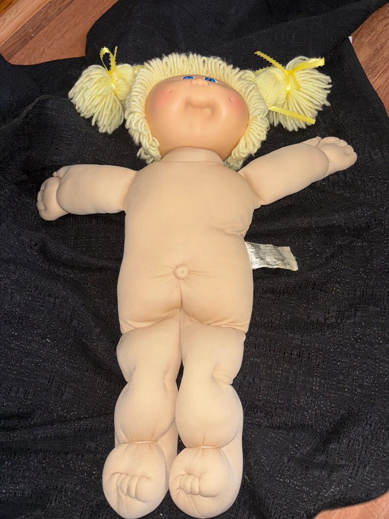 K&ouml;nnte beinhalten: Eine Vintage Cabbage Patch Kid Puppe mit einem weichen Stoffk&ouml;rper und einem Vinylkopf. Die Puppe hat blonde Zopf-Z&ouml;pfe, die mit gelben B&auml;ndern gebunden sind, blaue Augen und rosige Wangen. Die Puppe hat einen neutralen Hautton und einen Knopf am Po.