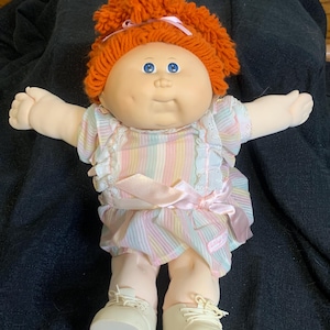 Op de afbeelding: Een Cabbage Patch Kid pop met rood garen haar, die een roze en wit gestreepte jurk draagt met een roze lint en witte kantversiering. De pop draagt witte schoenen met veters.