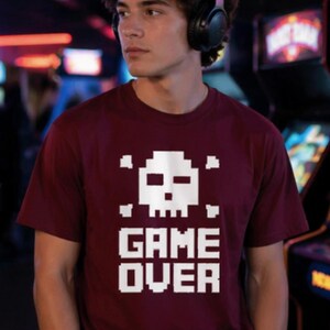 Pode incluir: Camiseta bord&ocirc; com um desenho de caveira e ossos cruzados pixelizados em branco. As palavras "GAME OVER" s&atilde;o impressas em letras brancas em bloco abaixo. A camiseta &eacute; de manga curta.