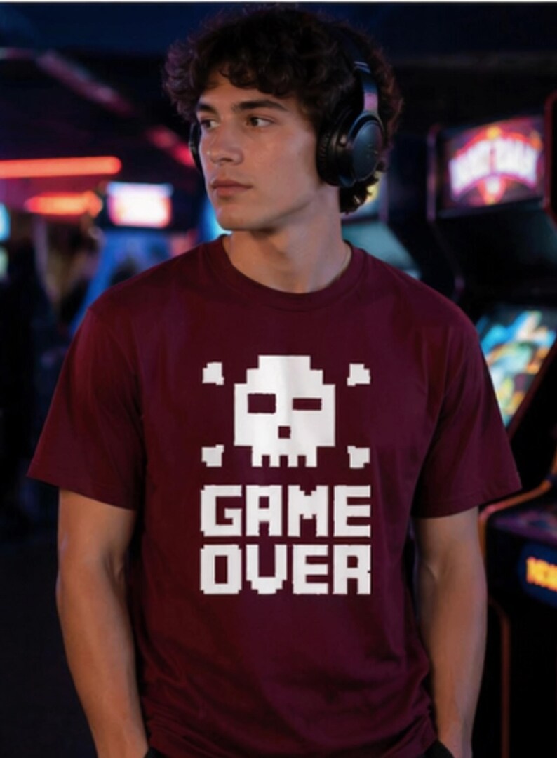 Pode incluir: Camiseta bord&ocirc; com um desenho de caveira e ossos cruzados pixelizados em branco. As palavras "GAME OVER" s&atilde;o impressas em letras brancas em bloco abaixo. A camiseta &eacute; de manga curta.