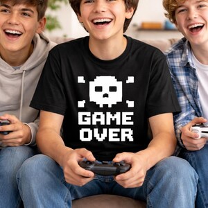 Pode incluir: Uma camiseta preta com uma caveira pixelizada e as palavras "GAME OVER" em branco. A camiseta &eacute; usada por uma pessoa segurando um controle de videogame, cercada por outras duas pessoas, todas sorrindo.