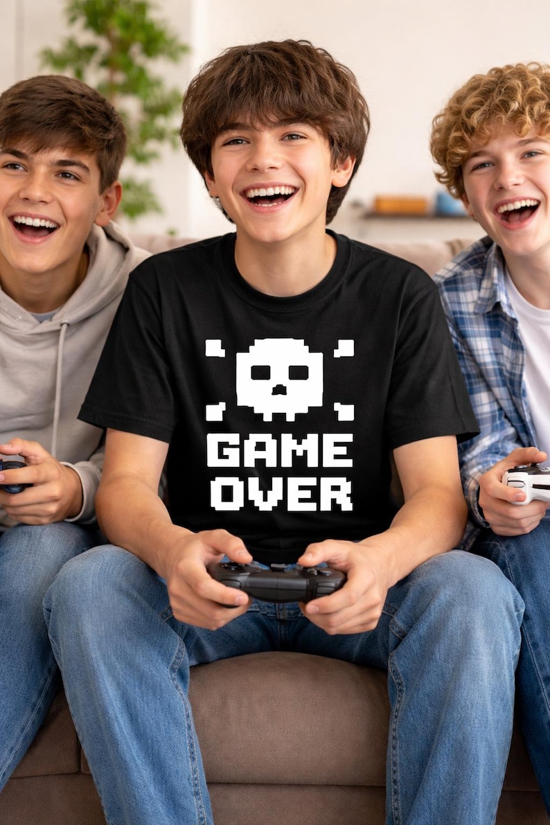 Pode incluir: Uma camiseta preta com uma caveira pixelizada e as palavras "GAME OVER" em branco. A camiseta &eacute; usada por uma pessoa segurando um controle de videogame, cercada por outras duas pessoas, todas sorrindo.