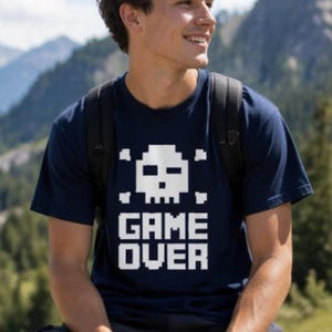 Pode incluir: Camiseta azul marinho com um design de caveira e ossos cruzados pixelizados em branco e a frase "GAME OVER". A camiseta &eacute; feita de um material macio e &eacute; adequada para roupas casuais.