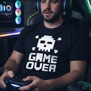 Pode incluir: Uma pessoa vestindo uma camiseta preta com uma caveira pixelada e as palavras "GAME OVER" em branco. Ele est&aacute; usando um fone de ouvido e segurando um controle de jogo, com um computador e um teclado ao fundo.