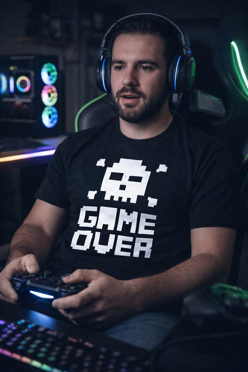 Pode incluir: Uma pessoa vestindo uma camiseta preta com uma caveira pixelada e as palavras "GAME OVER" em branco. Ele est&aacute; usando um fone de ouvido e segurando um controle de jogo, com um computador e um teclado ao fundo.