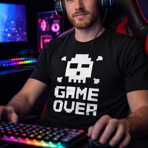 Pode incluir: Camiseta preta com um desenho pixelizado de caveira e ossos cruzados acima das palavras brancas "GAME OVER". A pessoa est&aacute; usando fones de ouvido e usando um teclado e mouse, sugerindo um tema de jogo.