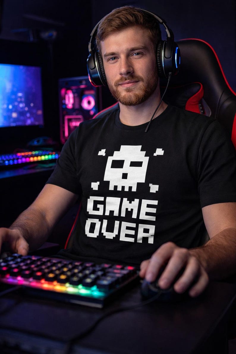 Pode incluir: Camiseta preta com um desenho pixelizado de caveira e ossos cruzados acima das palavras brancas "GAME OVER". A pessoa est&aacute; usando fones de ouvido e usando um teclado e mouse, sugerindo um tema de jogo.