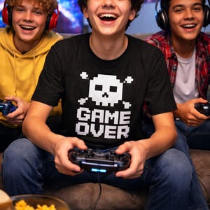 Pode incluir: Tr&ecirc;s jovens sorriem enquanto jogam videojogos. A pessoa do centro usa uma camiseta preta com um desenho pixelizado de caveira e ossos cruzados e as palavras "GAME OVER". Cada pessoa segura um controle de jogo.