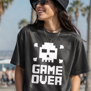 Pode incluir: Camiseta cinza escuro com um design de caveira e ossos cruzados pixelizados brancos acima das palavras "GAME OVER" em letras brancas. A pessoa est&aacute; usando um chap&eacute;u de balde preto e &oacute;culos de sol.