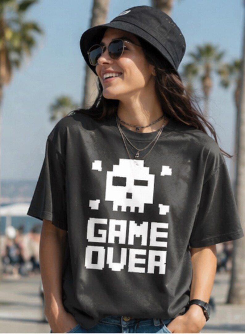 Pode incluir: Camiseta cinza escuro com um design de caveira e ossos cruzados pixelizados brancos acima das palavras "GAME OVER" em letras brancas. A pessoa est&aacute; usando um chap&eacute;u de balde preto e &oacute;culos de sol.