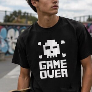 Pode incluir: Camiseta preta com um design de caveira e ossos cruzados pixelados em branco e a frase "GAME OVER". A camiseta de manga curta &eacute; usada por uma pessoa que segura um skate.