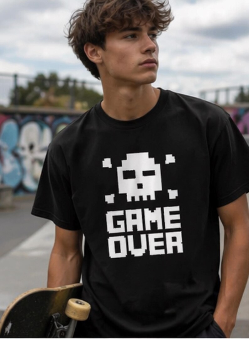 Pode incluir: Camiseta preta com um design de caveira e ossos cruzados pixelados em branco e a frase "GAME OVER". A camiseta de manga curta &eacute; usada por uma pessoa que segura um skate.