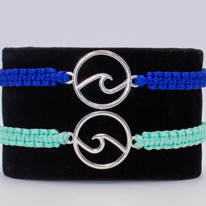 Puede incluir: Dos pulseras de macramé con un dije de plata en forma de ola. Las pulseras son azul y verde. El texto "AVAILABLE IN MORE COLORS!" está en la parte inferior de la imagen.