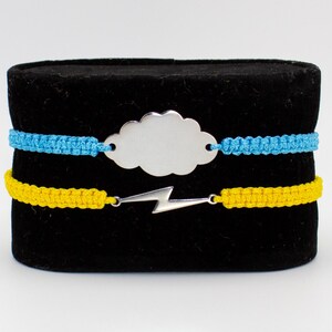 May include: Two friendship bracelets with silver charms. The top bracelet is blue and features a cloud charm. The bottom bracelet is yellow and features a lightning bolt charm.  "Macramuse Creations" is printed above the bracelets.