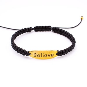 Puede incluir: Pulsera de macramé negra con un dije ovalado dorado que dice "Believe". La pulsera tiene nudos corredizos ajustables y cuentas doradas en los extremos. La pulsera se muestra sobre un fondo blanco.