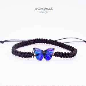 May include: Black macrame bracelet featuring a blue butterfly charm. The butterfly has a silver-colored border and a vibrant blue design. The text "AVAILABLE IN 13 COLORS!" is displayed in red.