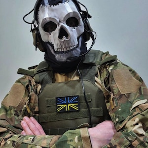Op de afbeelding: Een persoon met een schedelmasker, een headset en een camouflage militair uniform. Het masker is wit en grijs, met zwarte oogkassen en een gedetailleerd schedelontwerp. Het uniform omvat een groen vest met een patch met een blauw, geel en zwart ontwerp.