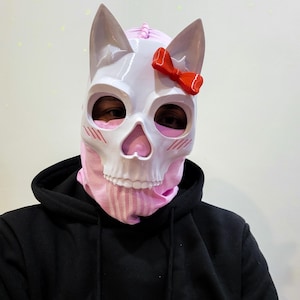 May include: A white skull mask with cat ears and a red bow, worn over a pink balaclava. The mask has eye and nose openings, and blush marks are painted on the cheeks. The person is wearing a black hooded sweatshirt.