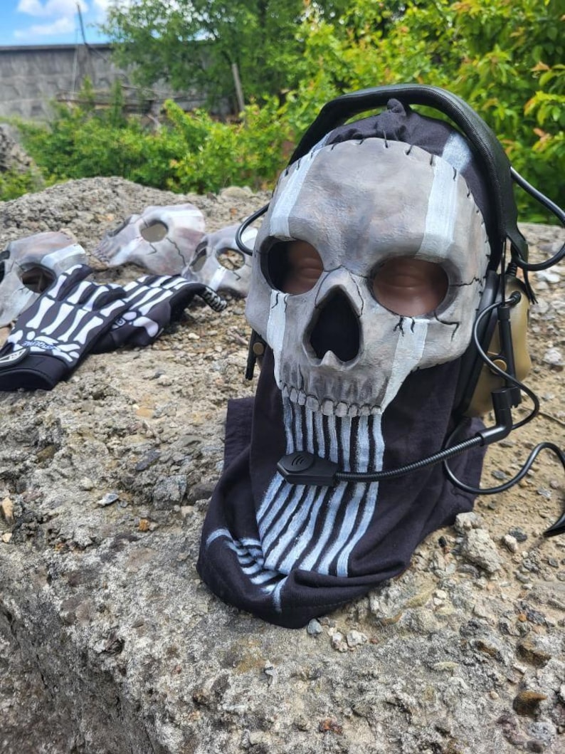 以下が含まれることがあります： 白いスケルトンデザインの黒いTシャツを着たスカルマスク。マスクはグレーと白で、黒い目の穴と黒いヘッドセットが付いています。他のマスクとスケルトングローブが背景にあります。