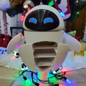Può includere: Una statuetta di robot bianco con un volto nero e occhi blu, che assomiglia al personaggio Eve del film Wall-E. Il robot ha scomparti aperti ed è circondato da una serie di luci natalizie colorate. Un albero di Natale è sullo sfondo.