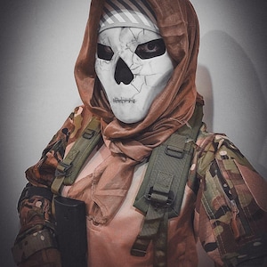 Peut inclure: Une personne portant un masque de crâne blanc avec une capuche marron et une veste de camouflage. Le masque a des orbites noires et une bouche cousue. La veste a des sangles vertes et un objet noir attaché.