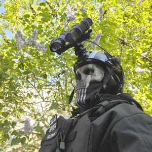 以下が含まれることがあります： 暗視ゴーグルとスカルをテーマにした顔覆いを装着したタクティカルヘルメット。ユニオンジャックのパッチと「Ultra Ghost」の文字が入った黒いタクティカルベストを着用。背景には緑の葉と青い空が写っています。