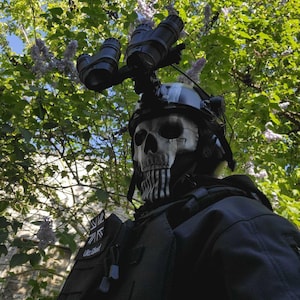 以下が含まれることがあります： 黒いタクティカルヘルメットに暗視ゴーグルとスカルマスク。マスクは黒いアクセントが付いた白いスカルデザインです。その人は「UK Ghost」のパッチが付いた黒いタクティカルベストを着ています。
